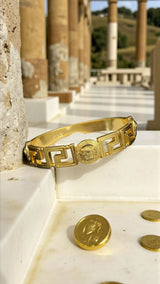 Medusa Aeterna Bracelet. Bold. Timeless. Captivating. This striking cuff features the iconic Medusa medallion centered between the legendary Greek Key motif. Crafted from durable stainless steel and richly plated in 21k gold.